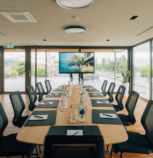 Lichtdurchfluteter Seminarraum mit großflächigen Fenstern, modernem Mobiliar und Aussicht ins Grüne. Der große Konferenztisch ist stilvoll gedeckt, mit Getränken, Schreibmaterial und Präsentationsbildschirm. Ideal für Meetings und Workshops in inspirierender Umgebung am Thunersee. Moderner Seminarraum mit Panoramablick