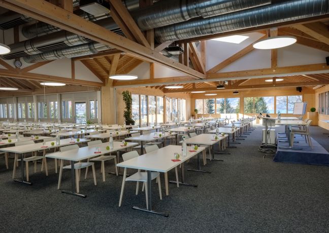 Moderner Tagungsraum mit viel Tageslicht, Holzarchitektur und langen Tischen – ideal für Seminare und Firmenanlässe am Thunersee. ©Seaside Spiez Heller Seminarraum mit Holzträgern und Reihenbestuhlung in der Thunersee-Region