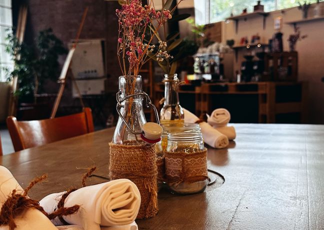 Detailaufnahme eines gedeckten Tisches mit Blumen, Kerzen und Servietten – warme, rustikale Atmosphäre für Business-Events in der Region Thun-Thunersee. ©Frachtraum Thun Stilvoll gedeckter Holztisch mit Vase und Stoffservietten in einer Eventlocation am Thunersee