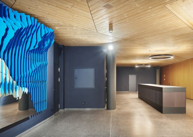 Lichtdurchflutete Lobby mit grosser Reception, warmen Holzflächen und markanter blauer Wandinstallation – stilvoller Empfang für Business-Events. ©Gemeinde Spiez Moderner Empfangsbereich mit Holzdecke und blauer Kunstinstallation in Spiez