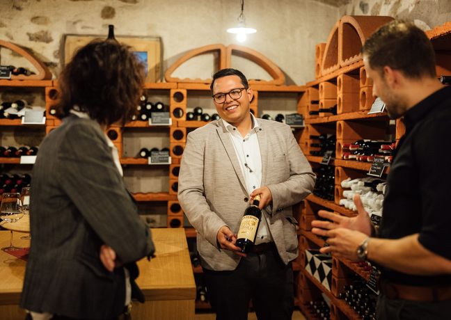 Drei Personen unterhalten sich entspannt in einem stilvollen Weinkeller in der Region Thunersee. Im Mittelpunkt steht eine Weinpräsentation vor rustikalen Weinregalen. Ideal als exklusive Event-Location für Schweizer Unternehmen und internationale Geschäftstreffen. ©Schützen Geselliges Beisammensein im Weinkeller