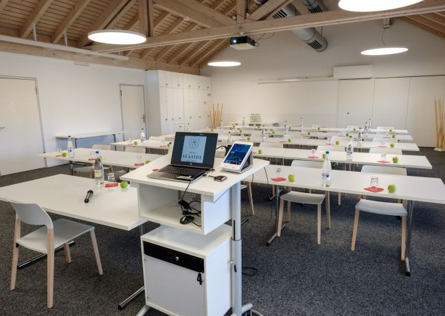 Heller Seminarraum mit U-Form-Bestuhlung, Screen und Moderationspult – ideal für Workshops, Meetings und Schulungen in der Thun-Thunersee Region. ©Seaside Spiez Seminarraum mit U-Form-Bestuhlung und Moderationspult in Spiez