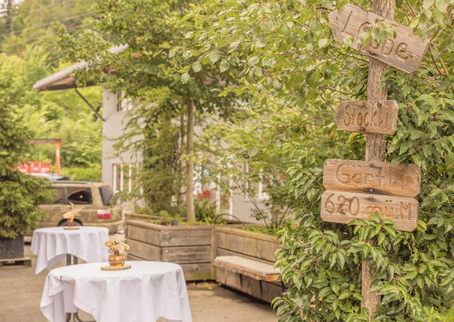 Heller Gartenhof mit zwei Stehtischen, Holzwegweiser und viel Grün – ideal für Networking und Apéro bei Firmenanlässen in der Thunersee-Region. ©Loube Spiez Stehtische im grünen Innenhof mit Wegweiser-Schildern für Outdoor-Apéro