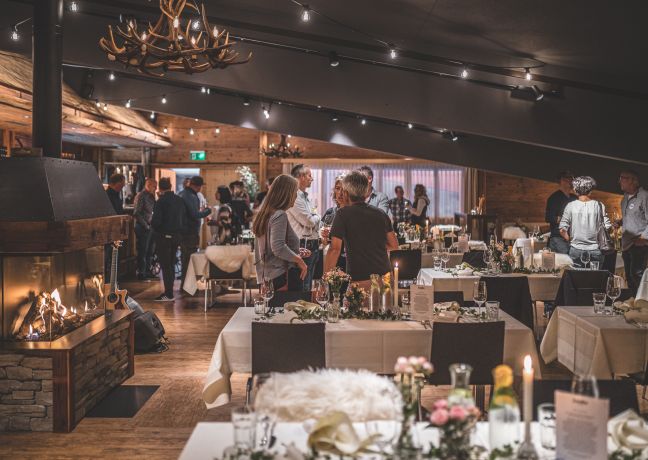 Gemütliche Eventlocation mit warmem Licht, Kronleuchter und Kaminfeuer: gedeckte Tafeln, Blumenarrangements und Gäste im Austausch schaffen eine einladende Atmosphäre. Ideal für Firmenfeiern, Incentives oder Networking-Events am Thunersee in der Schweiz – professionell, charmant und inspirierend. ©Loube Spiez Stilvolles Dinner-Event am Thunersee