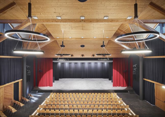 Blick von hinten in einen Holzsaal mit Bühne, roten Vorhängen und Reihenbestuhlung – ideal für Tagungen und Firmenanlässe in der Thunersee-Region. ©Gemeinde Spiez Moderner Veranstaltungssaal mit Bühne, Reihenbestuhlung und Ringlichtern in Thun