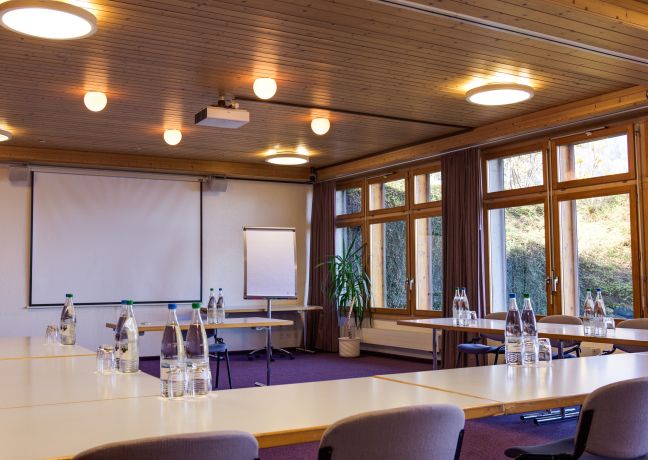 Moderner, lichtdurchfluteter Seminarraum mit U-Form-Bestuhlung, Beamer und grosser Leinwand. Die Fensterfront bietet Blick ins Grüne und schafft eine ruhige Arbeitsatmosphäre – ideal für Workshops, Meetings und Weiterbildungen von KMU, Verbänden und internationalen Organisationen in der Region Thunersee. ©Meielisalp Heller Seminarraum am Thunersee