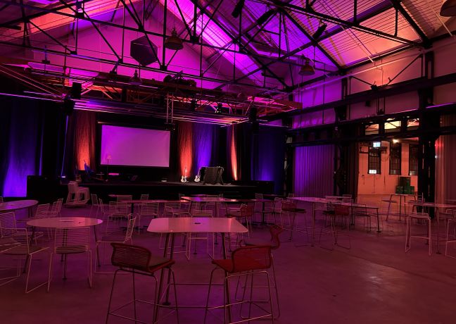 Spacious industrial event venue with round tables, a stage and a projection screen – atmospherically lit for meetings and corporate events. ©Lady Evelyn Event hall with a stage and screen, atmospheric purple lighting and seating areas