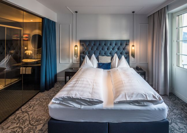 Hell eingerichtetes Hotelzimmer mit Boxspringbett, Glaswand zum Bad und Fensterblick – ideal für Business-Gäste und MICE-Aufenthalte in Thun. ©Parkhotel, Gunten Modernes Doppelzimmer mit Boxspringbett und blauer Polsterwand am Thunersee