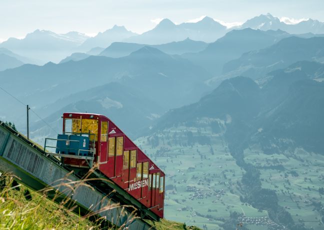 Eine rote Bergbahn fährt steil bergauf; im Hintergrund liegen Thunersee-Region und Alpenpanorama in morgendlicher Klarheit. ©Niesen Rote Schmalspurbahn fährt am Berghang mit Blick auf die Alpen