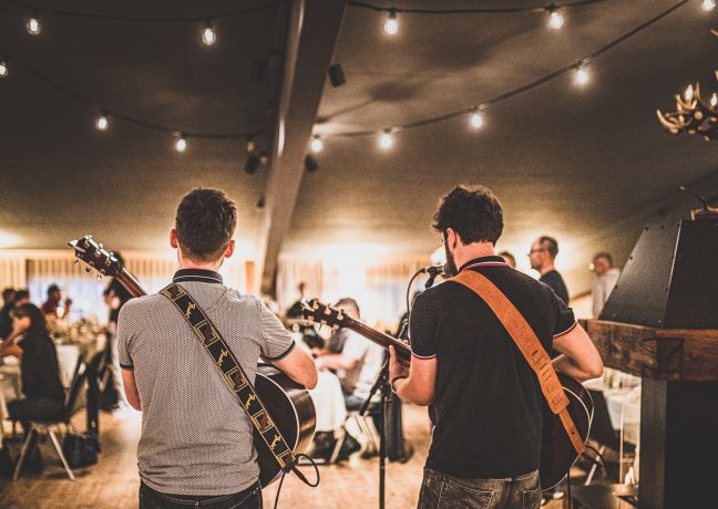 Zwei Musiker mit Gitarren sorgen für Stimmung im beleuchteten Saal – ideal für MICE-Events und Abendprogramme in der Region Thun-Thunersee. ©Loube Spiez Live-Band spielt bei einem Firmenevent im Festzelt am Thunersee
