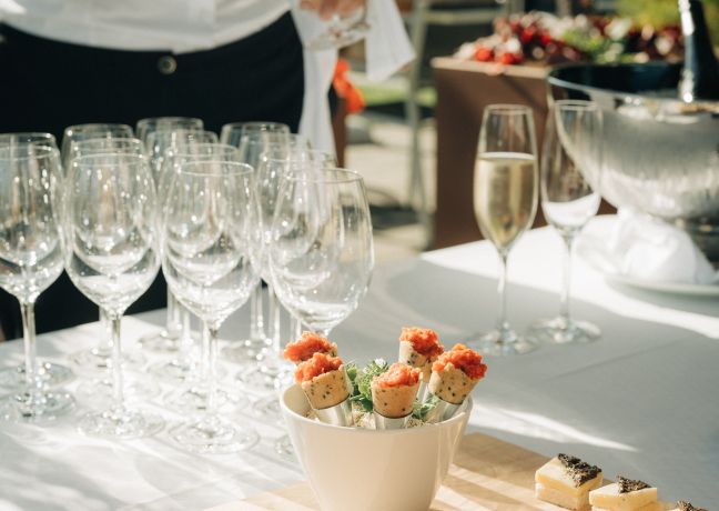 Ein stilvoll gedeckter Tisch im Freien präsentiert Sektgläser, die frisch eingeschenkt werden. Feine Canapés und süße Häppchen sorgen für Genussmomente bei Ihrem Seminar in Thun. Die sonnige Atmosphäre lädt zum entspannten Netzwerken und Genießen ein. ©Deltapark Vitalresort, Thun Edle Seminarpause mit Sekt und Häppchen