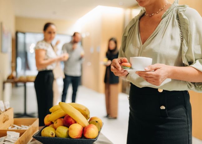 Während einer Seminarpause genießen Teilnehmende frisches Obst und Kaffee. Im Vordergrund steht eine Schale mit Äpfeln und Bananen, während die entspannte Atmosphäre eines modernen Veranstaltungsraums spürbar ist. Ideal für produktive Meetings in Thun und am Thunersee. Seminarpause mit frischem Obst