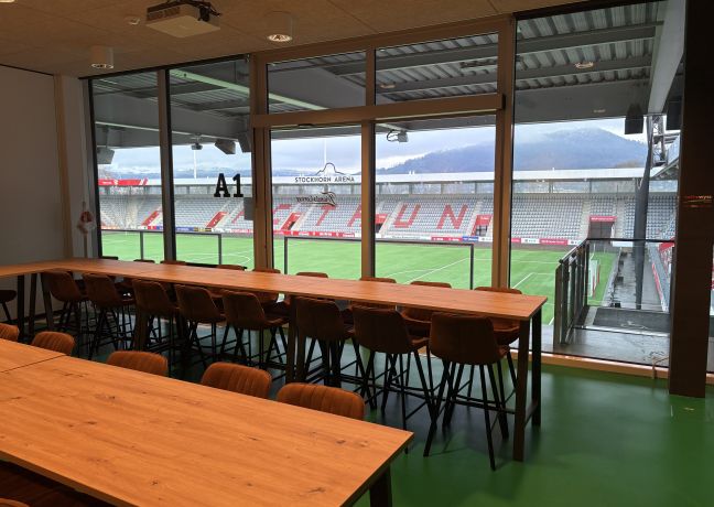 Modernes Meeting-Setting mit Holztischen und Reihenbestuhlung; durch die Glasfront fällt der Blick direkt auf das Spielfeld im Stadion Thun. ©Stockhorn Arena Seminarraum mit Panoramafenster und Blick ins Stadion Thun