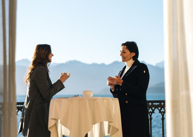 Two businesswomen enjoy a relaxed coffee break at a round standing table on a sunny terrace with unobstructed views of sparkling Lake Thun and the Alps. Ideal for inspiring conversations and networking in stylish surroundings. Business coffee break with a view of Lake Thun