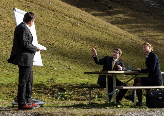 Drei Geschäftsleute halten ein Meeting an einem Holztisch inmitten der Natur mit sanften Hügeln im Hintergrund. Ein Flipchart steht bereit, während eine Person präsentiert und die anderen aufmerksam zuhören. Ideal für kreative Tagungen und Teamevents in entspannter Atmosphäre am Thunersee. ©Stockhorn Geschäftsmeeting im Freien am Thunersee