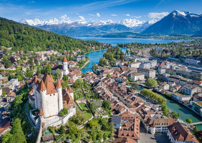 Beeindruckende Luftaufnahme von Thun, im Vordergrund das historische Schloss und die Schlosskirche. Der türkisfarbene Fluss schlängelt sich durch die Altstadt, im Hintergrund glitzert der Thunersee vor der imposanten Kulisse der Berner Alpen. Perfekte Kulisse für inspirierende Firmenevents in der Schweiz. ©Thun-Thunersee Tourismus Luftaufnahme von Thun mit Schloss und Alpenpanorama