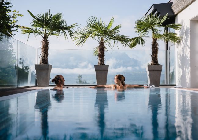 Zwei Personen genießen entspannte Momente im modernen Außenpool mit Blick auf die Alpenkulisse und den Thunersee. Palmen und Glasbalustraden sorgen für ein exklusives, mediterranes Ambiente – ideal für inspirierende Geschäftsmeetings oder Teamevents in einzigartiger Schweizer Umgebung. ©Solbad Entspannende Poolatmosphäre mit Alpenblick