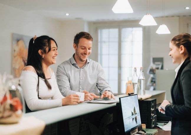 Freundlicher Empfang am Front Desk: Zwei Gäste besprechen Unterlagen mit der Gastgeberin – ideal für Meetings und Business-Events am Thunersee. ©Hotel Krone Business-Gäste beim Check-in an einer Hotelrezeption im Thunersee-Meeting-Setting