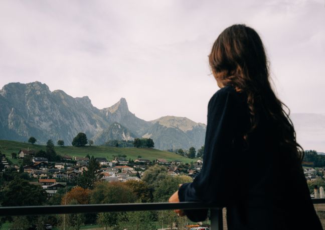 Eine Frau genießt die malerische Aussicht von einem Hotelbalkon auf ein idyllisches Schweizer Dorf und die imposanten Berge der Berner Alpen. Die entspannte Atmosphäre und das weite Panorama laden zum Verweilen ein und machen den Aufenthalt in Thun besonders. ©Deltapark Aussicht vom Hotelbalkon auf die Berner Alpen