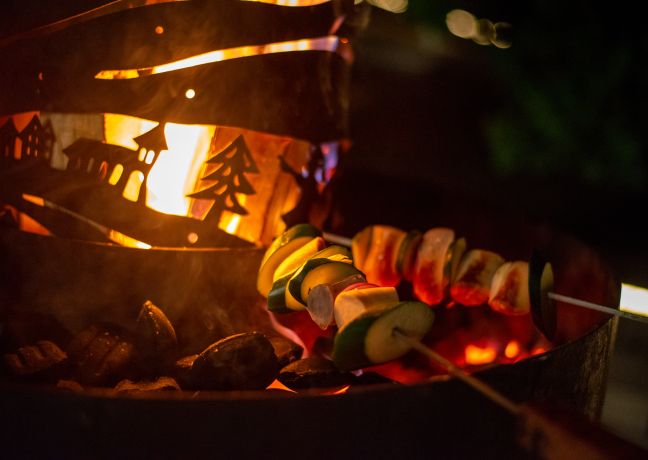 Ein stimmungsvolles Bild von buntem Grillgemüse auf Spießen, das über einem offenen Lagerfeuer mit dekorativem Windschutz im Alpenstil gegrillt wird. Perfekt für Teamevents und gesellige Firmengrillabende in der Region Thunersee. Die warme Beleuchtung sorgt für ein einladendes Ambiente. ©Seaside Spiez Gemütliches Grillen am Lagerfeuer