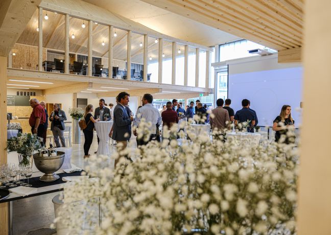 Teilnehmende vernetzen sich beim Apéro in hellem, modernem Innenraum mit Galerie, grossen Fenstern und Blumenarrangement im Vordergrund. ©Westhalle Thun Business-Apéro im modernen Eventraum mit Stehbuffet und Gästen