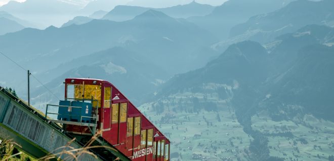 A red narrow-gauge railway runs along the mountainside with views of the Alps